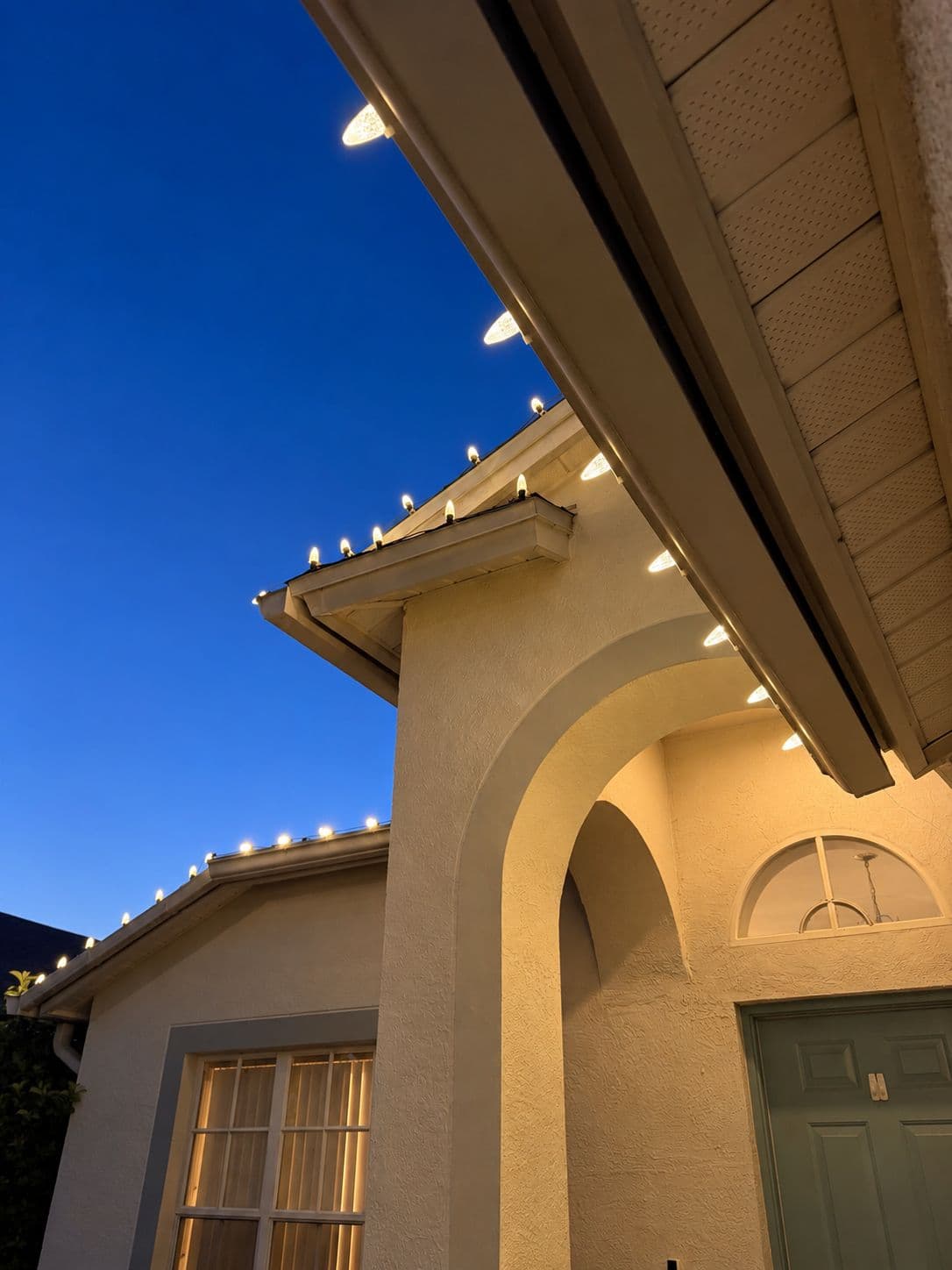 Looking up at an arched entry at dusk, with C9 bulbs clipped evenly along the gutter and gable
