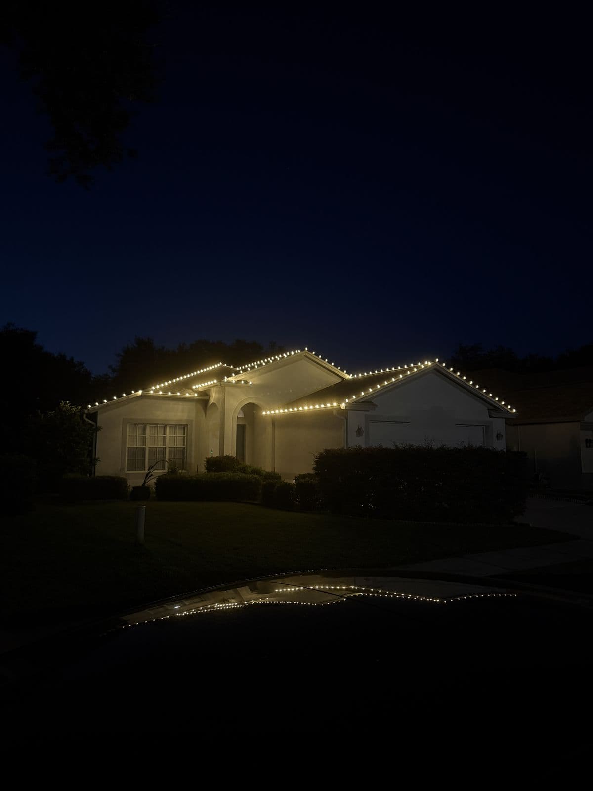 A home at night with every roofline outlined in warm white lights, reflected in a car hood in the foreground