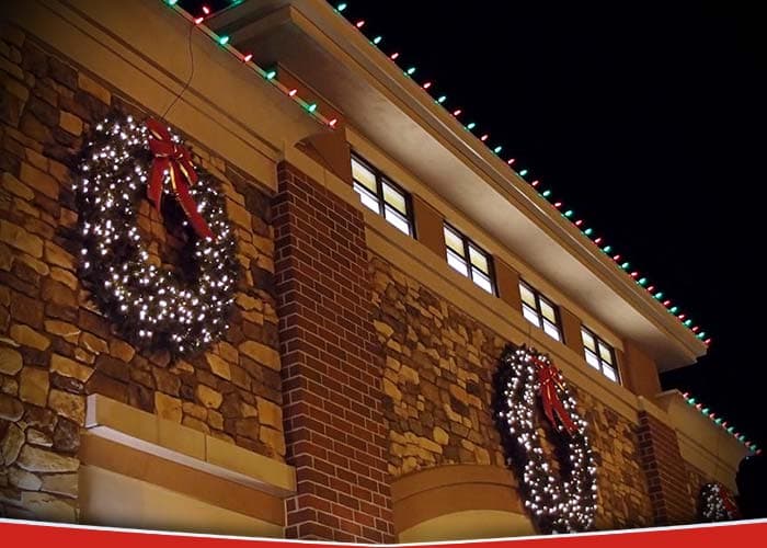 A brick and stone commercial building at night edged with red and green C9 lights, with large lit wreaths and red bows mounted on the façade